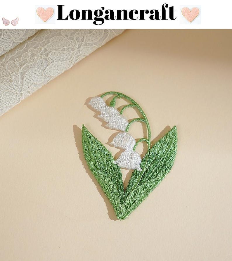May include: Embroidered lily of the valley patch with white flowers and green leaves. The patch is in the shape of a heart. The text "Longancraft" is above the patch.
