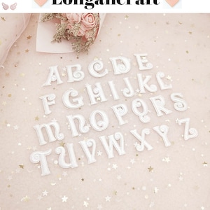 Może przedstawiać: Białe, haftowane litery alfabetu ułożone na jasnoróżowej powierzchni z konfetti w kształcie gwiazd. Litery są w ozdobnej czcionce. Słowo "Longancraft" znajduje się na górze obrazu.