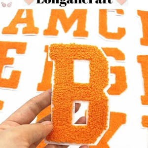 Pode incluir: Patches de letras de feltro laranja em maiúsculas. As letras são A, M, C, E, B, G, I e K. As letras estão sobre um fundo branco.