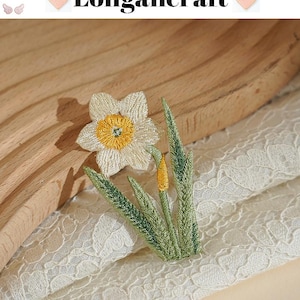 May include: Embroidered daffodil patch with a white flower, yellow center, and green leaves. The patch is on a cream-colored lace fabric. The text "Longancraft" is above the flower.