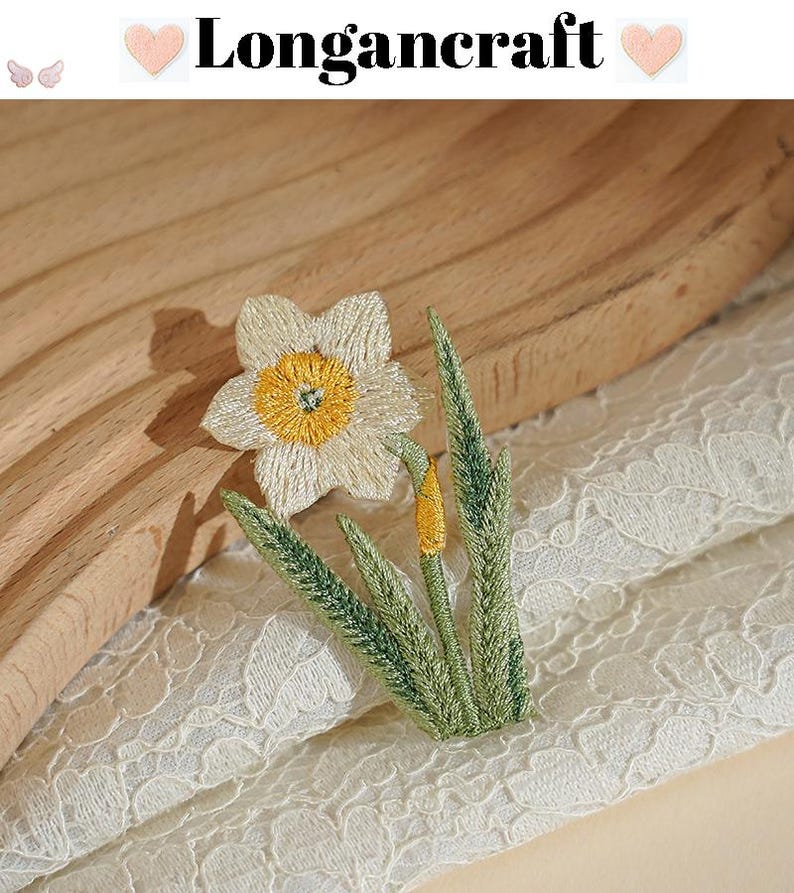 May include: Embroidered daffodil patch with a white flower, yellow center, and green leaves. The patch is on a cream-colored lace fabric. The text "Longancraft" is above the flower.