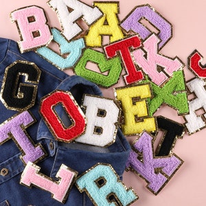 May include: Assortment of colorful felt letter patches with gold glitter trim. The patches are in various colors, including pink, blue, green, yellow, red, white, and black. The patches are arranged in a pile on a pink surface.