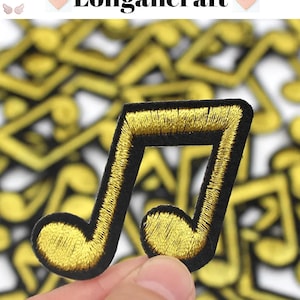 May include: A close-up of a gold musical note iron-on patch. The patch is embroidered with gold thread on a black fabric background. The patch is held in a hand.