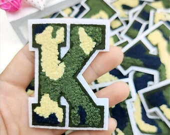May include: A green, yellow, and black camouflage fabric letter K patch. The patch is surrounded by a white border and has a fuzzy texture.
