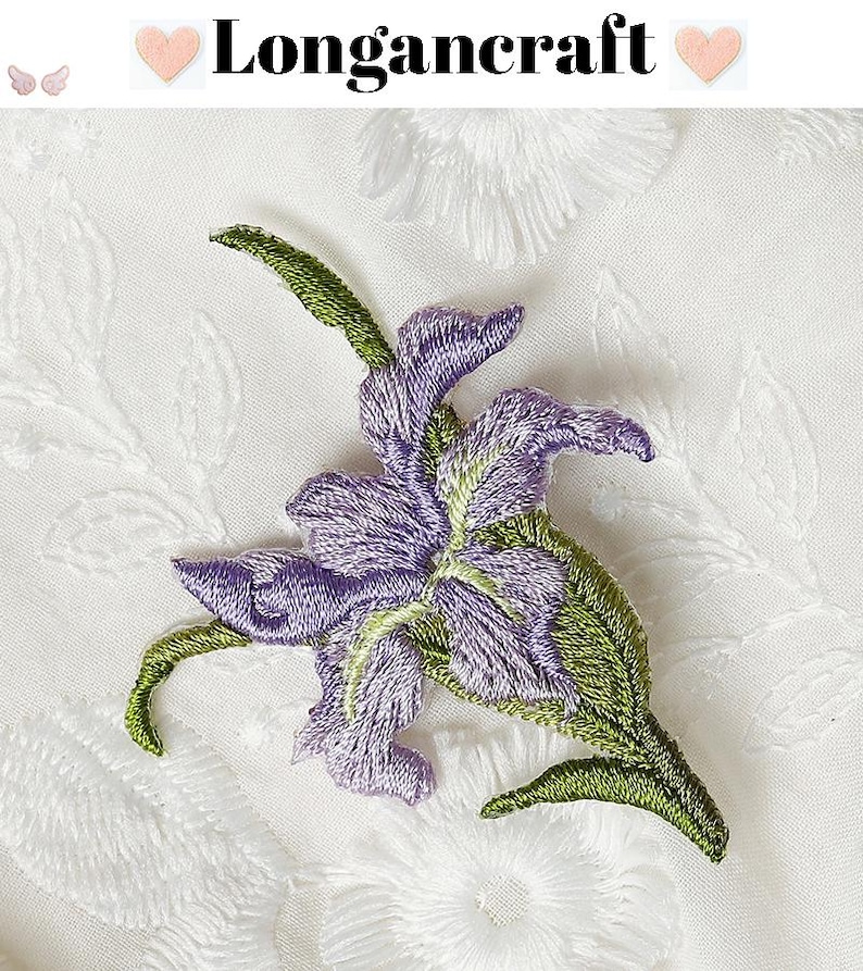 May include: Embroidered purple and green floral applique. The applique features a detailed design of a flower with purple petals and green leaves. The background is white with a subtle floral pattern. The text "Longancraft" is at the top.