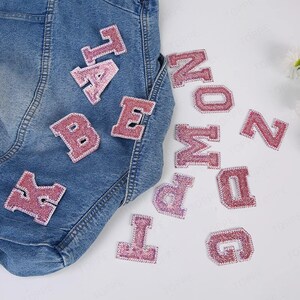 May include: Pink iron-on letters of the alphabet, A, B, E, G, I, K, M, N, O, P, T, Z. The letters are on a white background with a denim jacket in the background.
