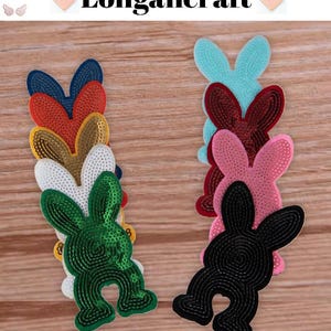 May include: A selection of colourful bunny-shaped fabric patches. The patches are in various colours, including blue, red, gold, white, green, pink, and black. The patches are arranged on a wooden surface. The text "Longancraft" is displayed at the top.
