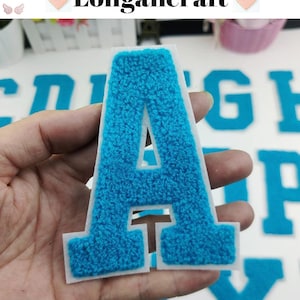 May include: A blue felt letter A with a white outline. The letter is on a white background and is being held by a hand.