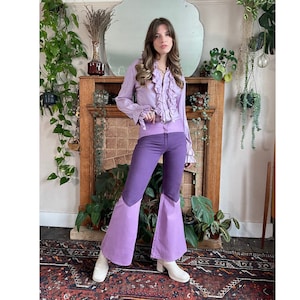 Könnte beinhalten: Eine Frau trägt eine lavendelfarbene Bluse mit Rüschen und eine violette und lavendelfarbene Schlaghose. Sie steht vor einem Kamin mit Pflanzen auf beiden Seiten.