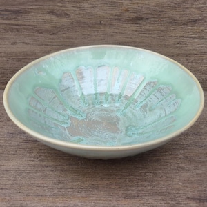 May include: A handmade ceramic bowl with a light green glaze and a unique sunburst pattern in the center. The bowl has a slightly oval shape and a cream-colored rim. The bowl is approximately 8 inches in diameter.