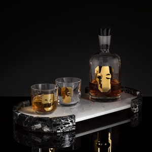 Könnte beinhalten: Ein Tablett aus schwarz-weißem Marmor mit goldfarbenem Rand hält eine klare Glaskaraffe und zwei Gläser. Die Karaffe und die Gläser sind mit goldenen Gesichtsdesigns verziert.