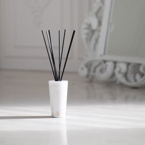 Op de afbeelding: Een witte keramische diffuser met zwarte rietjes in een witte kamer met een witte, sierlijke spiegel op de achtergrond.