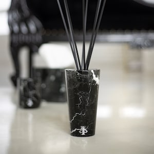 Puede incluir: Botella difusora de mármol negro con cañas negras y un logotipo blanco. La botella está sobre una superficie blanca.