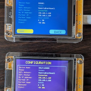 May include: Two small electronic devices with clear plastic cases display configuration information on their screens. The screens show text including "Device Name," "WiFi Network," and IP addresses. One screen displays "ROGUE1" and "Ocean," the other "GOLD-LEADER" and "Sunset."
