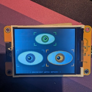 May include: A rectangular electronic display module with an orange circuit board frame. The screen shows three stylized blue eye icons within digital targeting brackets against a grid background, with the text OMNISCIENT WATCH NETWORK 3 at the bottom.