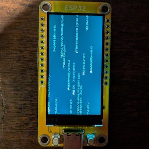 May include: A yellow ESP32 development board with a small screen displaying blue text. The board has multiple connection pins and a USB-C port. The text on the screen appears to be in Japanese characters.