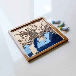 Pode incluir: Um mapa de madeira de uma cidade com uma incrusta&ccedil;&atilde;o de resina azul que representa a &aacute;gua. O mapa &eacute; emoldurado em uma moldura de madeira natural.