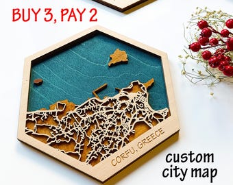 May include: A wooden custom city map of Corfu, Greece, in a hexagonal frame. The map is laser-cut with intricate details, set against a teal background. Text on the map reads "CORFU, GREECE" and "custom city map".