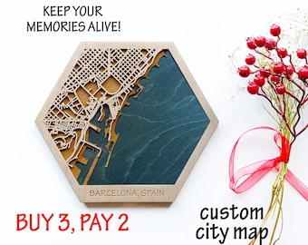 May include: A hexagonal wooden map of Barcelona, Spain, with a detailed laser-cut design. The map features a teal blue background and is accompanied by a bouquet of red berries and the text "KEEP YOUR MEMORIES ALIVE!"