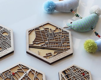 May include: Four hexagonal wooden coasters with laser-cut city maps. Each coaster features a different city and the text "OUR FIRST HOME" with the city and state name. The coasters are arranged on a white surface with a blue and white bird mobile hanging above.