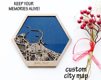 May include: A wooden hexagonal map of Bari, Italy, with a blue background and a laser-cut city map. The text "Keep your memories alive!" and "Bari, Italy" are engraved on the map. The map is on a white surface with a red ribbon and a bouquet of red berries.