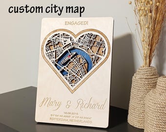 May include: A custom city map carved into a wooden plaque. The map, shaped like a heart, features a detailed street layout with a blue river. The text "ENGAGED!" is above the heart, and below it are the names "Mary & Richard", date, and coordinates. The words "custom city map" are at the top.