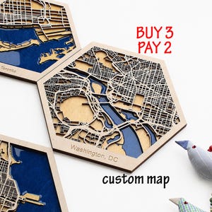 Puede incluir: Arte de pared hexagonal de madera con un mapa 3D en capas de Washington, DC, con detalles de agua azul y cuadrículas de calles en madera natural. El texto dice BUY 3 PAY 2 y custom map. La pieza incluye un diseño cortado con láser con profundidad.