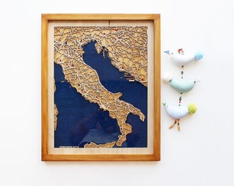 May include: Wooden framed map of Italy with intricate laser-cut details, showcasing the country's geography. The map features a blue background representing the sea and a light brown landmass. Decorative hanging birds with pom-poms are to the right.