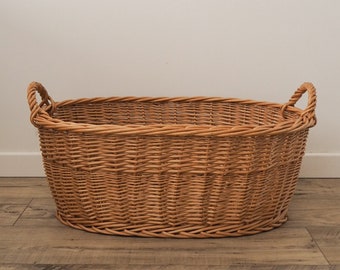 May include: A large, oval-shaped wicker basket with two handles. The basket is made of natural woven wicker and has a light brown color.