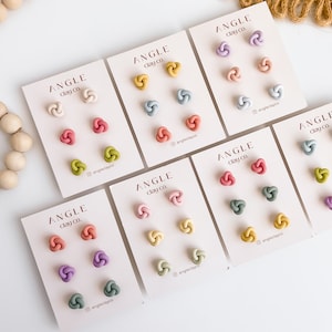 Op de afbeelding: Zes sets kleioorbellen in verschillende kleuren, waaronder roze, groen, blauw en geel. Elke set heeft drie oorbellen met een geknoopt ontwerp. De oorbellen worden gepresenteerd op witte kaartjes met de tekst "ANGLE Clay co."