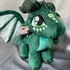 K&ouml;nnte beinhalten: Ein gr&uuml;ner Pl&uuml;schdrache mit gro&szlig;en Augen und einem hellgr&uuml;nen Bauch. Der Drache hat ein Paar Fl&uuml;gel und ein kleines Horn auf dem Kopf.