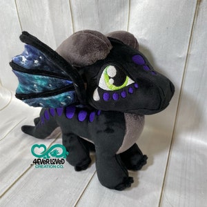 K&ouml;nnte beinhalten: Ein schwarzer und grauer Pl&uuml;schdrache mit violetten Akzenten und gr&uuml;nen Augen. Der Drache hat einen Fl&uuml;gel mit Galaxiemuster.