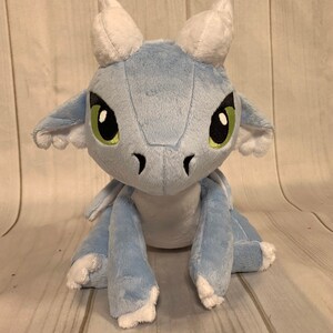 Puede incluir: Un drag&oacute;n de peluche azul claro con cuernos blancos y ojos verdes. El drag&oacute;n est&aacute; sentado sobre una superficie de madera.