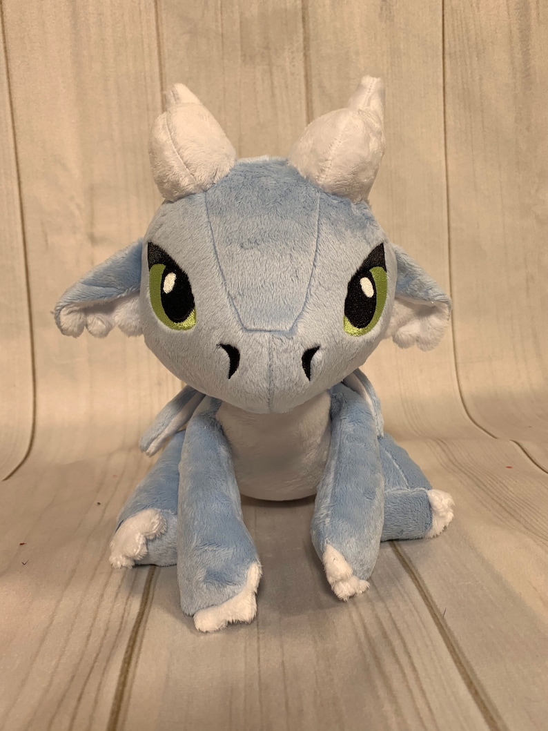 Puede incluir: Un drag&oacute;n de peluche azul claro con cuernos blancos y ojos verdes. El drag&oacute;n est&aacute; sentado sobre una superficie de madera.