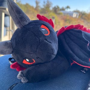 May include: A black dragon plush toy with red detailing on its wings and head. The dragon has orange-accented eyes. It rests on a blue surface, with a black strap attached.