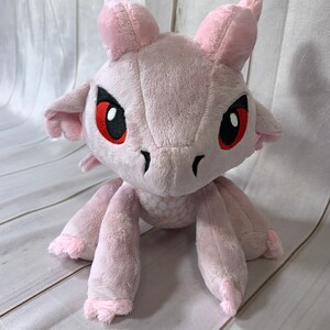 Puede incluir: Drag&oacute;n de peluche rosa con ojos rojos y escamas blancas. El drag&oacute;n est&aacute; sentado con las patas extendidas.