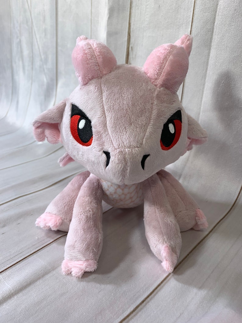 Puede incluir: Drag&oacute;n de peluche rosa con ojos rojos y escamas blancas. El drag&oacute;n est&aacute; sentado con las patas extendidas.