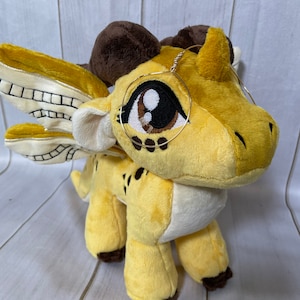 K&ouml;nnte beinhalten: Ein gelber Pl&uuml;schdrache mit braunen Akzenten und einem goldenen Drahtgestell-Brillenaccessoire. Der Drache hat ein kleines Horn auf dem Kopf und ein Paar Fl&uuml;gel.
