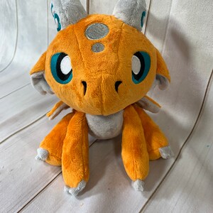 Puede incluir: Un drag&oacute;n de peluche naranja con cuernos y alas grises. El drag&oacute;n tiene grandes ojos azules y una mancha plateada en la cabeza.
