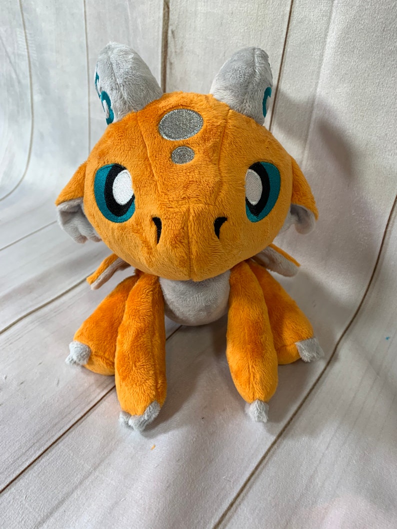 Puede incluir: Un drag&oacute;n de peluche naranja con cuernos y alas grises. El drag&oacute;n tiene grandes ojos azules y una mancha plateada en la cabeza.