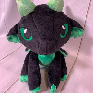 Puede incluir: Un drag&oacute;n de peluche negro y verde con grandes ojos verdes. El drag&oacute;n est&aacute; sentado sobre una superficie de madera blanca.