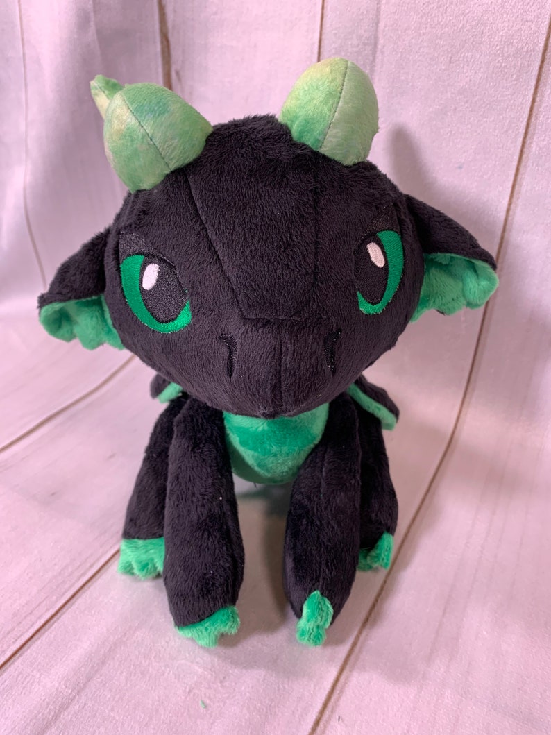 Puede incluir: Un drag&oacute;n de peluche negro y verde con grandes ojos verdes. El drag&oacute;n est&aacute; sentado sobre una superficie de madera blanca.