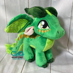 K&ouml;nnte beinhalten: Gr&uuml;ner Pl&uuml;schdrache mit gro&szlig;en Augen, orangefarbenen Flecken und blattf&ouml;rmigen Ohren. Der Drache steht auf einer wei&szlig;en Holzoberfl&auml;che. 4Ever Loved Creation Co. ist auf der Unterseite des Drachen aufgedruckt.