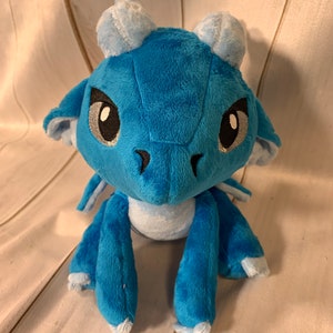 Puede incluir: Un drag&oacute;n de peluche azul con detalles blancos y ojos negros. El drag&oacute;n est&aacute; sentado con las piernas cruzadas y las alas dobladas detr&aacute;s de &eacute;l.