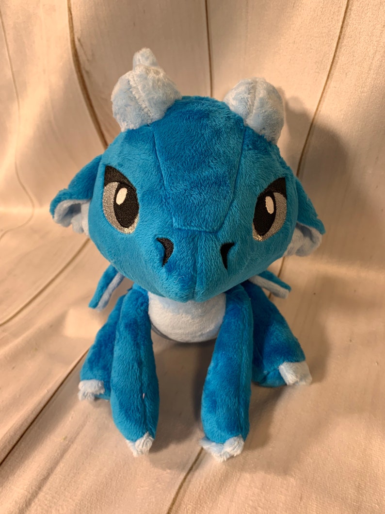 Puede incluir: Un drag&oacute;n de peluche azul con detalles blancos y ojos negros. El drag&oacute;n est&aacute; sentado con las piernas cruzadas y las alas dobladas detr&aacute;s de &eacute;l.