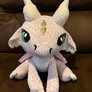 Puede incluir: Un drag&oacute;n de peluche rosa y blanco con detalles dorados. El drag&oacute;n tiene ojos verdes y un cuerpo blanco con manchas. Est&aacute; sentado sobre una superficie marr&oacute;n.