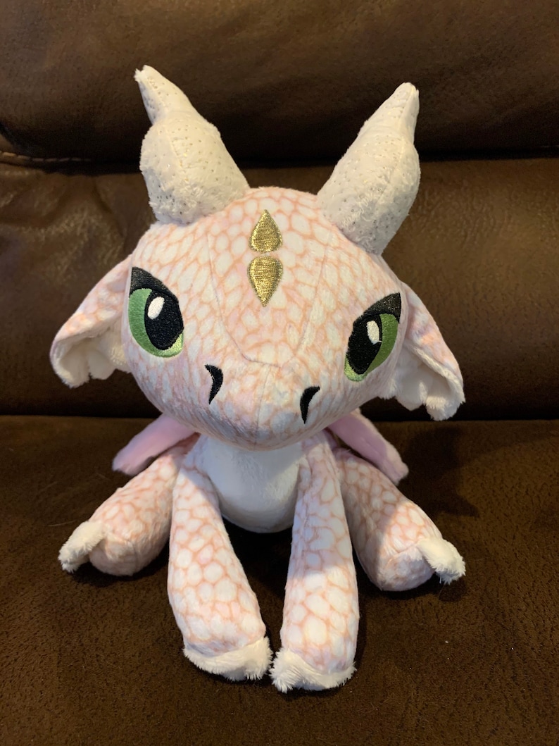Puede incluir: Un drag&oacute;n de peluche rosa y blanco con detalles dorados. El drag&oacute;n tiene ojos verdes y un cuerpo blanco con manchas. Est&aacute; sentado sobre una superficie marr&oacute;n.
