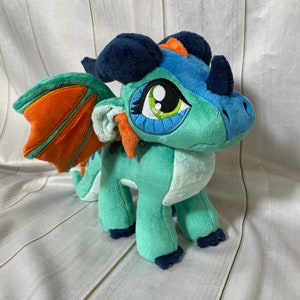 K&ouml;nnte beinhalten: Ein tealfarbener und orangefarbener Pl&uuml;schdrache mit blauen Augen und einem dunkelblauen Kamm. Der Drache hat einen kleinen orangenen Fl&uuml;gel auf der einen Seite und einen gr&ouml;&szlig;eren orangenen Fl&uuml;gel auf der anderen Seite.