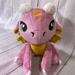 Puede incluir: Un drag&oacute;n de peluche rosa y amarillo con cuernos blancos y ojos verdes. El drag&oacute;n est&aacute; sentado sobre una superficie de madera blanca.