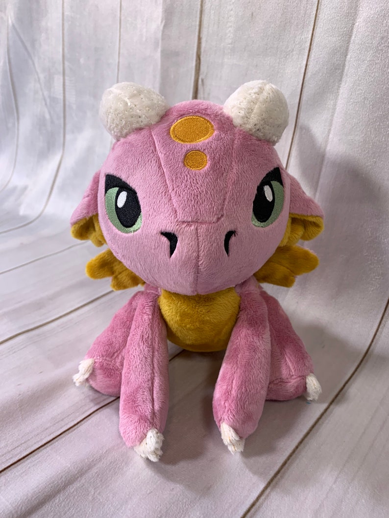 Puede incluir: Un drag&oacute;n de peluche rosa y amarillo con cuernos blancos y ojos verdes. El drag&oacute;n est&aacute; sentado sobre una superficie de madera blanca.